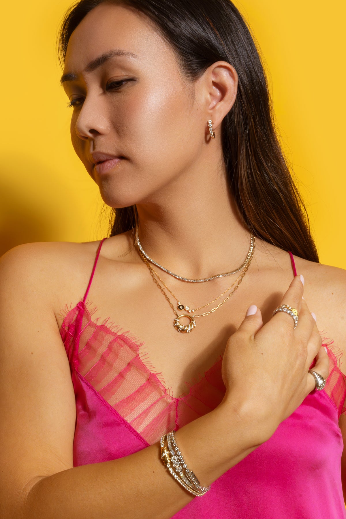 Model in a pink top wearing Petite Frites 18K gold and diamond jewelry including bracelets, necklaces, rings and earrings against a yellow background.