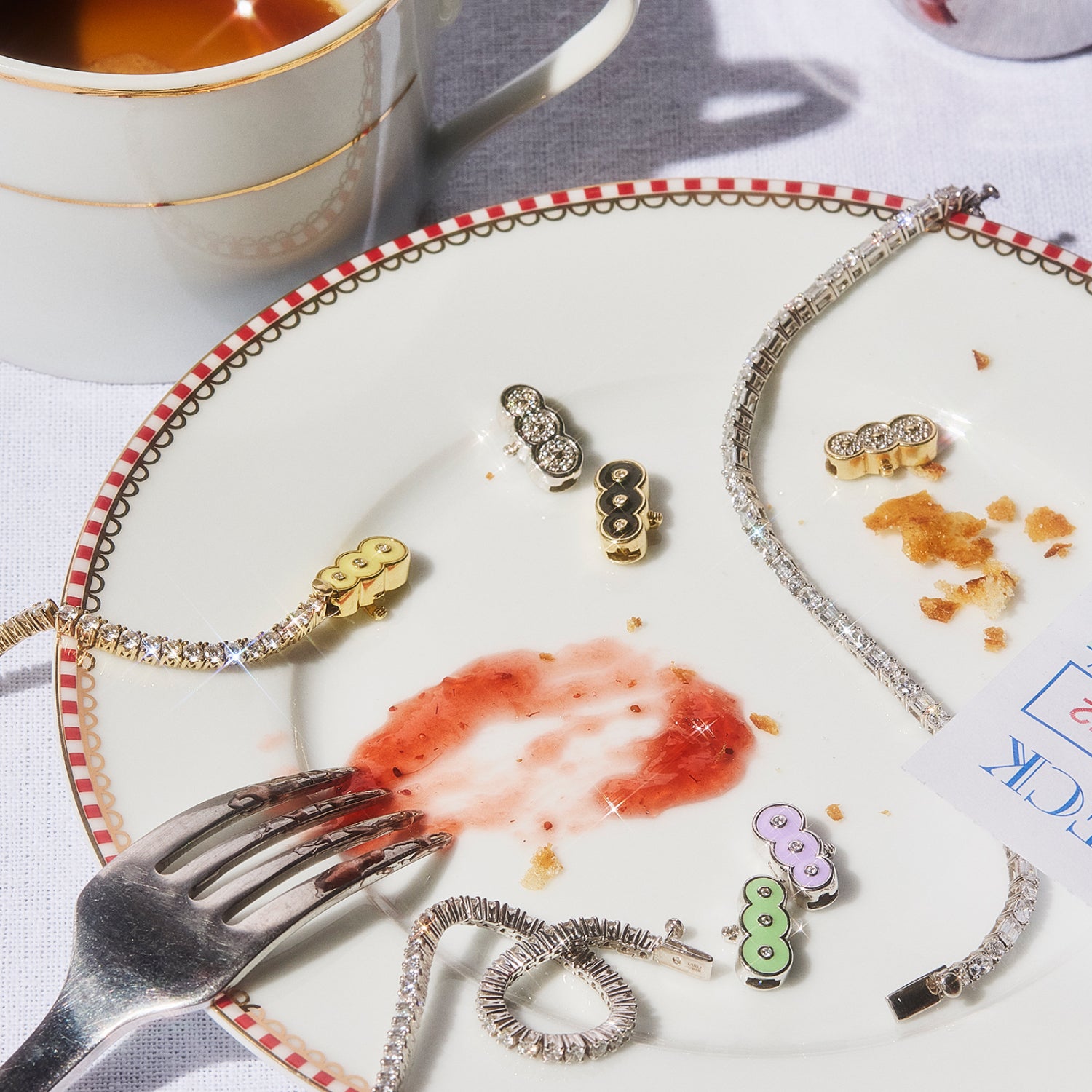 Elegant Petite Frites diamond jewelry, including bracelets and colorful clasps, displayed on a decorative plate with crumbs and a smear of jam. A tea cup sits nearby, evoking a playful yet sophisticated moment.