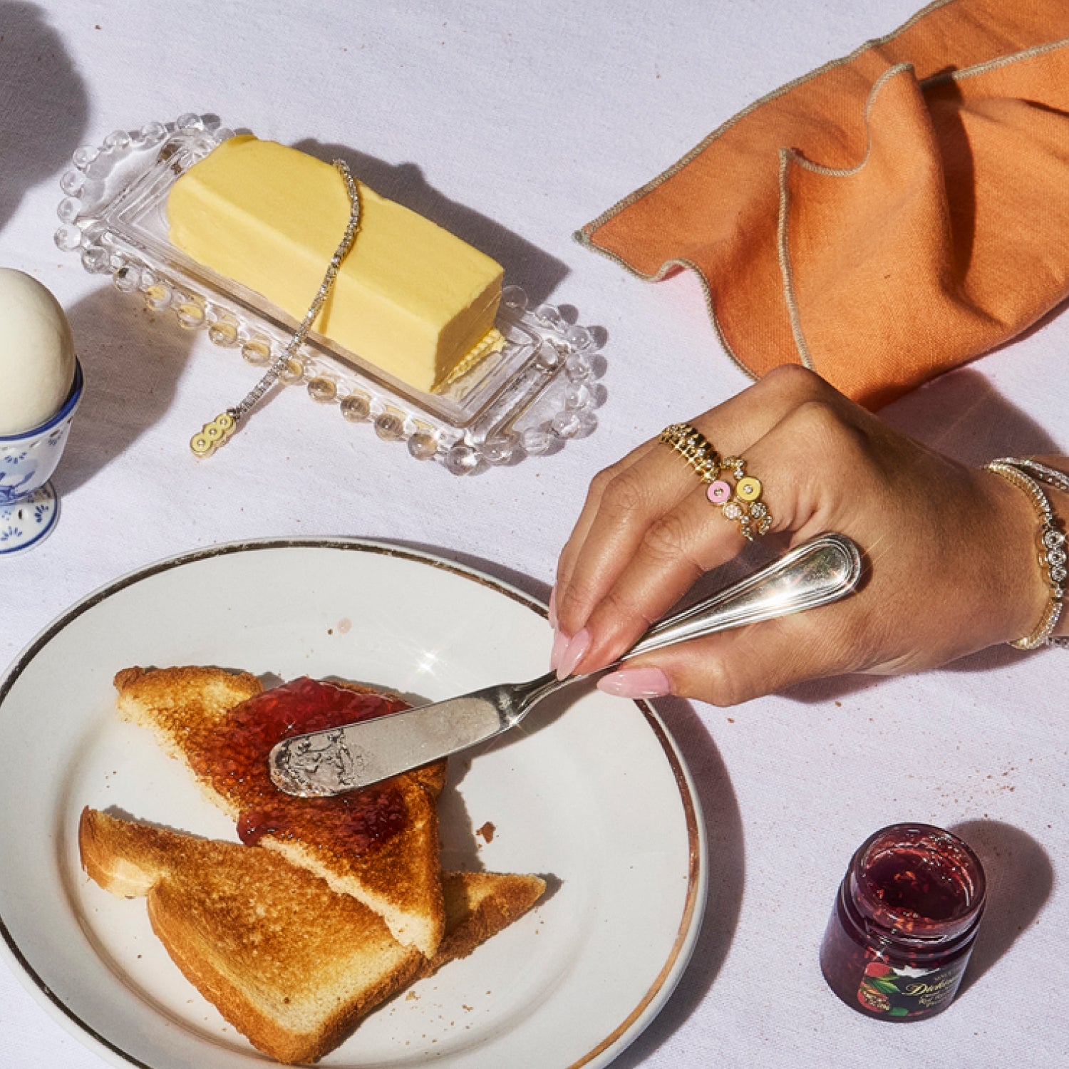 Hand adorned with Petite Frites fine diamond jewelry, including rings, and tennis bracelets, displayed on a white linen tablecloth with a plate of buttered toast and jams. The image highlights the playful, bold, and luxurious designs of Petite Frites’ collection.
