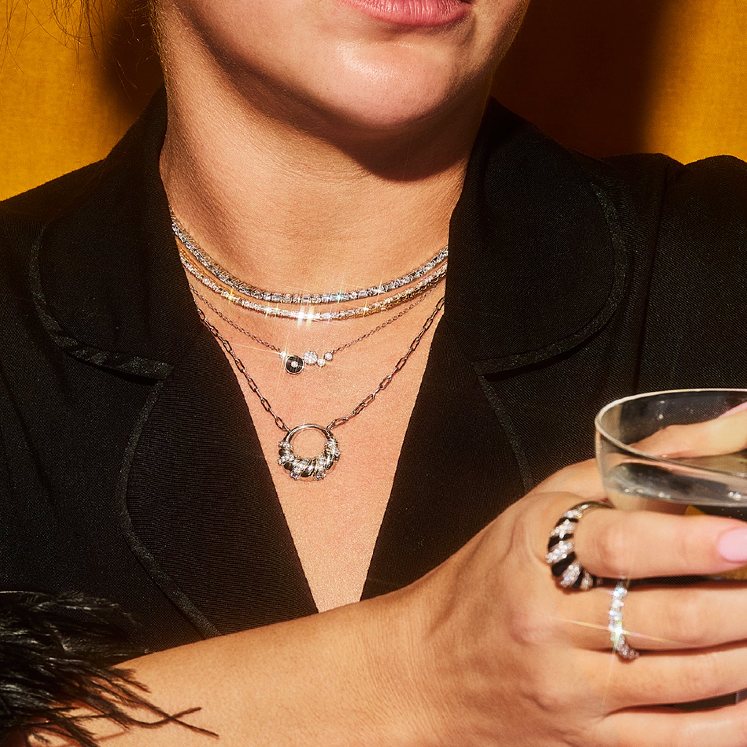 A woman wearing layered Petite Frites diamond necklaces and rings with a black blazer, raising her glass to cheers. The image highlights the playful, bold, and luxurious designs of Petite Frites’ collection.