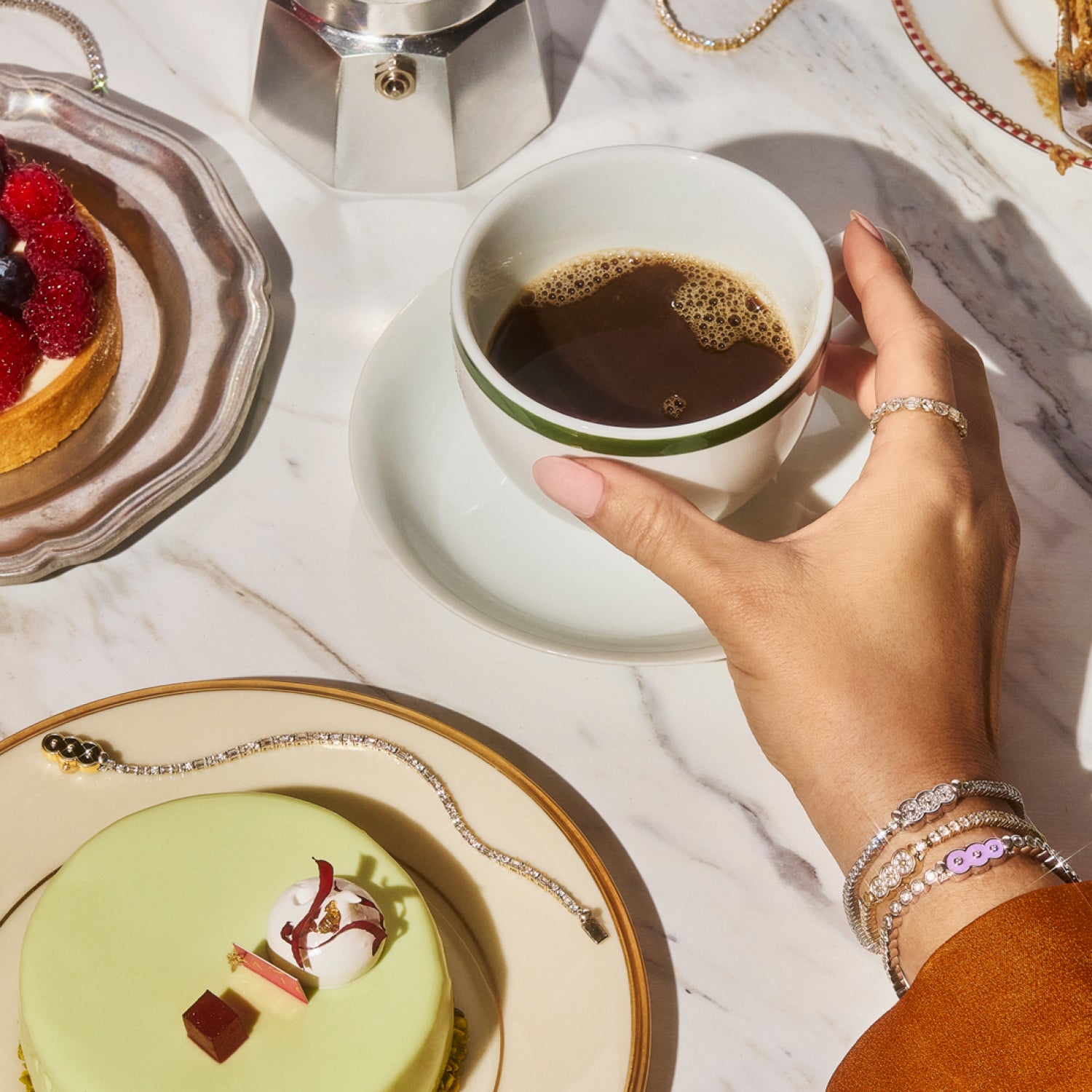 Hand adorned with Petite Frites fine diamond jewelry, including rings, tennis bracelets, displayed on a marble table alongside a cup of coffee and decadent dessert pastries. The image highlights the playful, bold, and luxurious designs of Petite Frites’ collection.