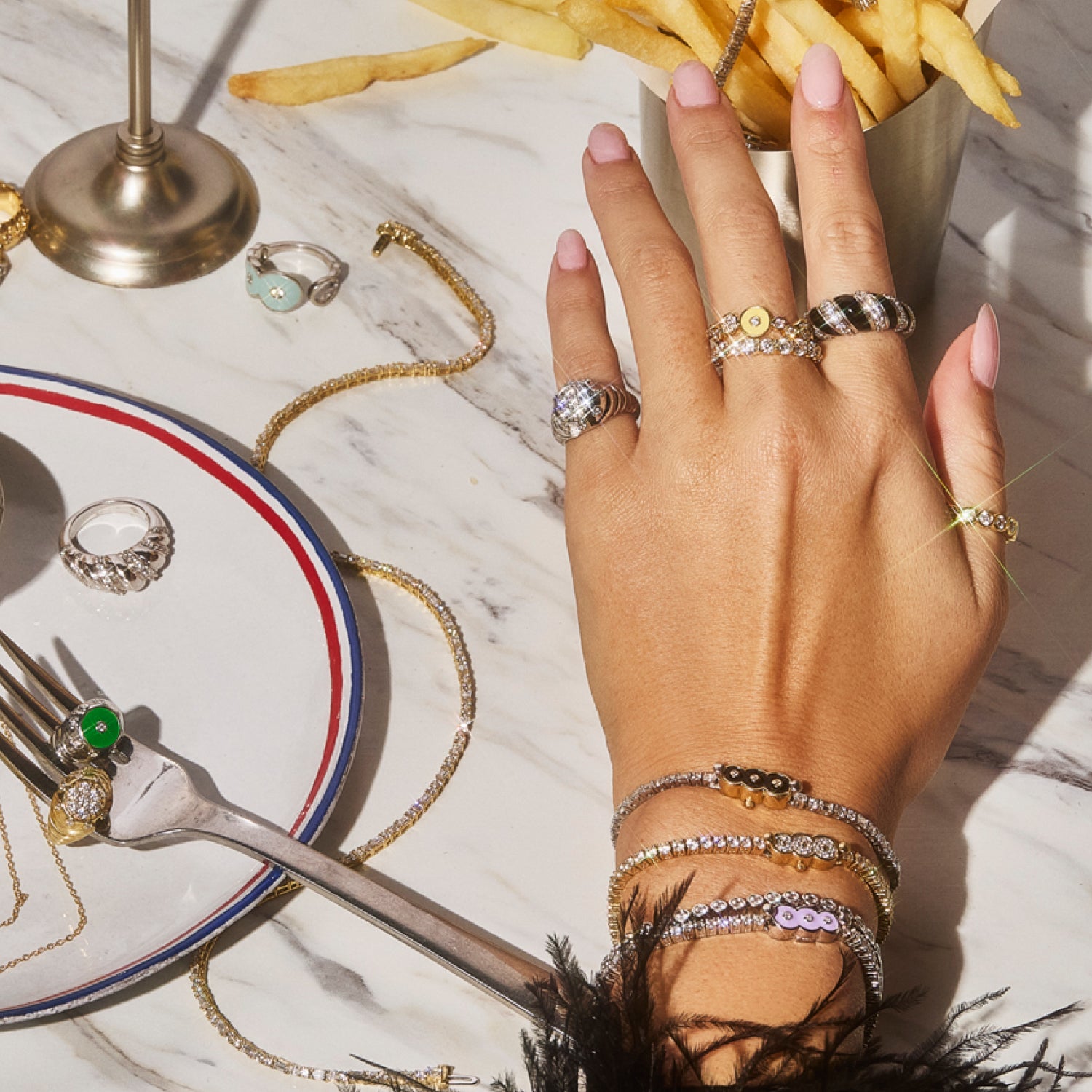 Hand adorned with Petite Frites fine diamond jewelry, including rings, bracelets, and necklaces, displayed on a marble table alongside a plate of fries and elegant tableware. The image highlights the playful, bold, and luxurious designs of Petite Frites’ collection.