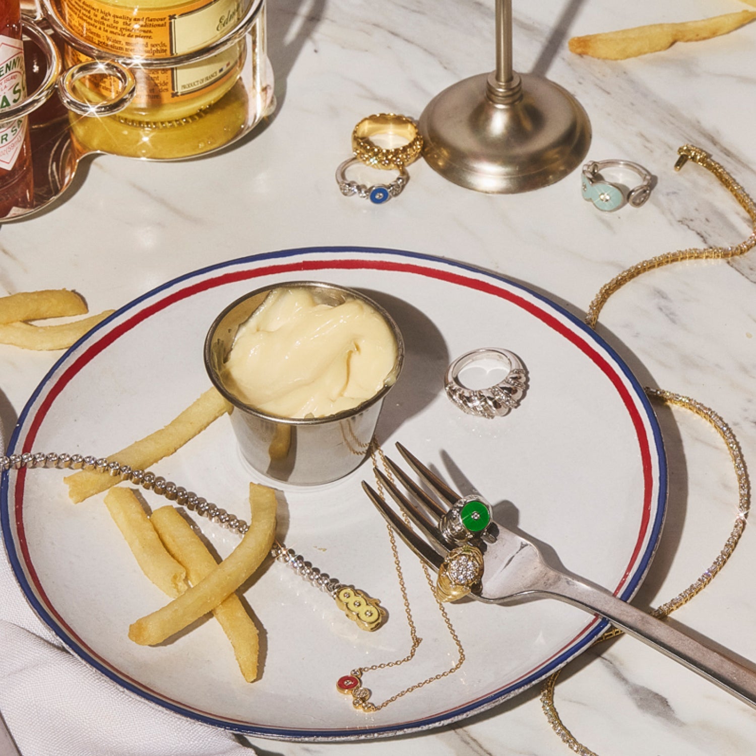 Petite Frites fine diamond jewelry, including bold rings and playful colorful designs, creatively styled on a fork and decorative plate against a marble tabletop, blending sophistication with whimsy