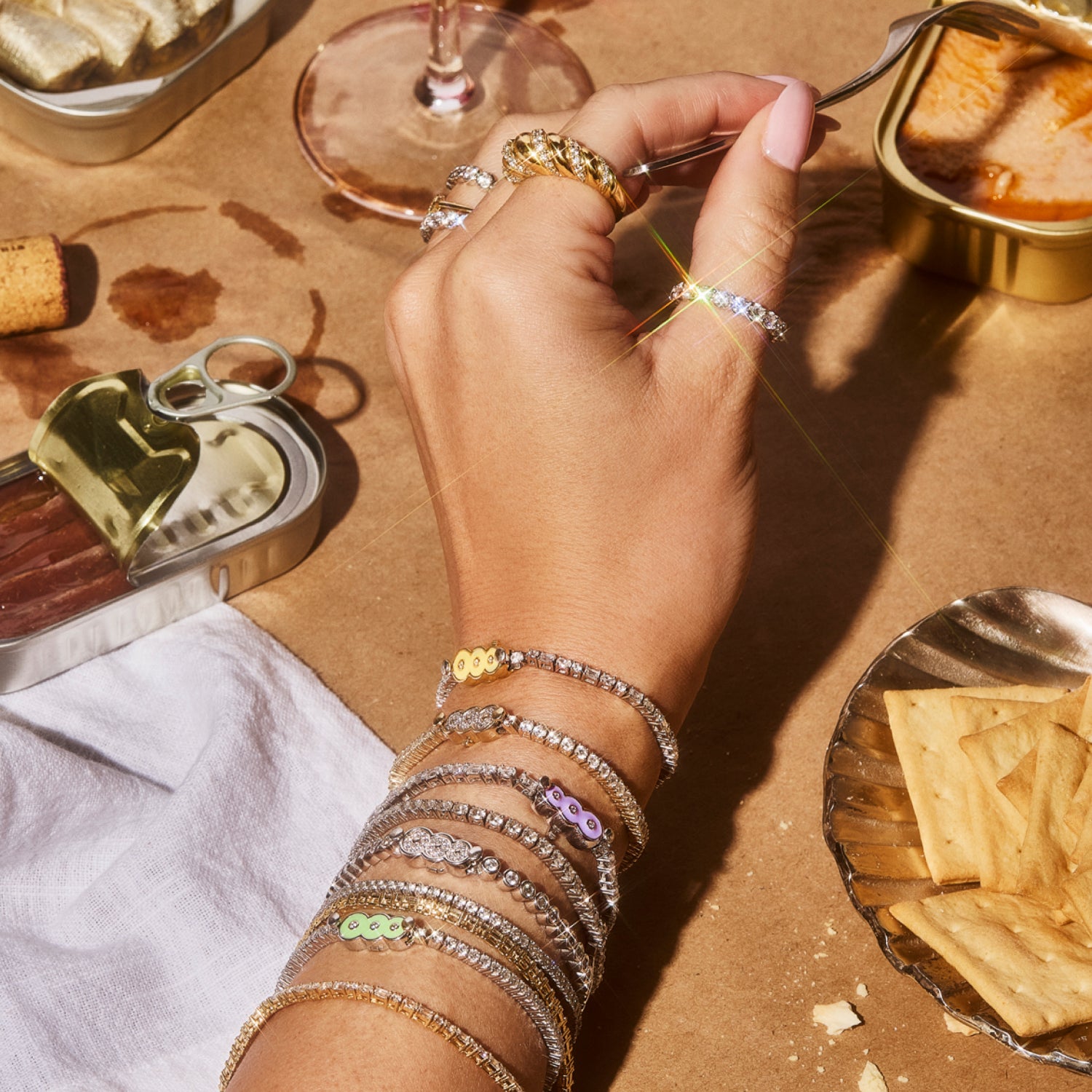 A hand and wrist adorned with multiple Petite Frites 18K white and yellow gold diamond tennis bracelets and diamond and gold rings reaching for a tin can of sardines.