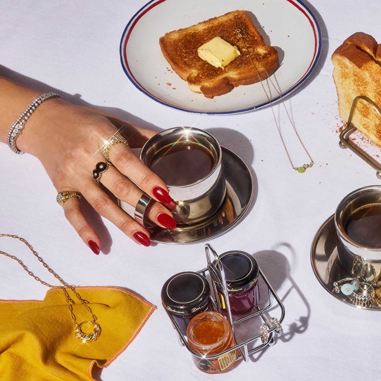 Hand adorned with Petite Frites fine diamond jewelry, including rings, and tennis bracelets, displayed on a linen table alongside a teacup, toast with butter and elegant tableware. The image highlights the playful, bold, and luxurious designs of Petite Frites’ collection.