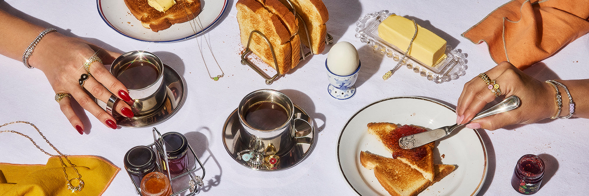 This image shows a breakfast spread of coffee, toast, butter and jam, with two female hands reaching for their respective coffee and toast dishes. The hands are adorned with Petite Frites 18k gold and carbon neutral lab grown diamond tennis bracelets and rings in white and yellow gold, with colorful enamel accents. On the table are more rings and necklaces placed across the food dishes.