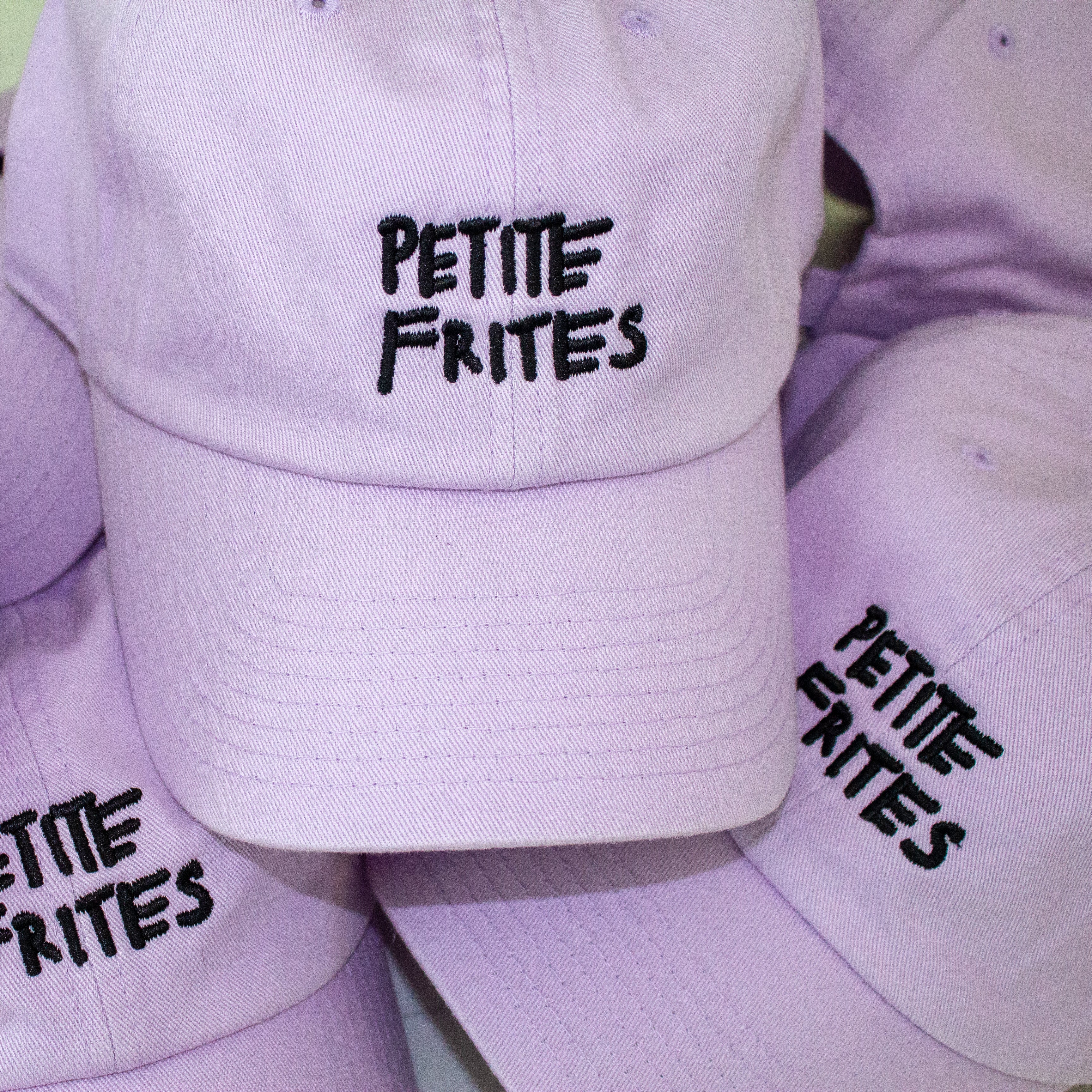 A pile of lavender baseball caps with the Petite Frites logo embroidered in black.
