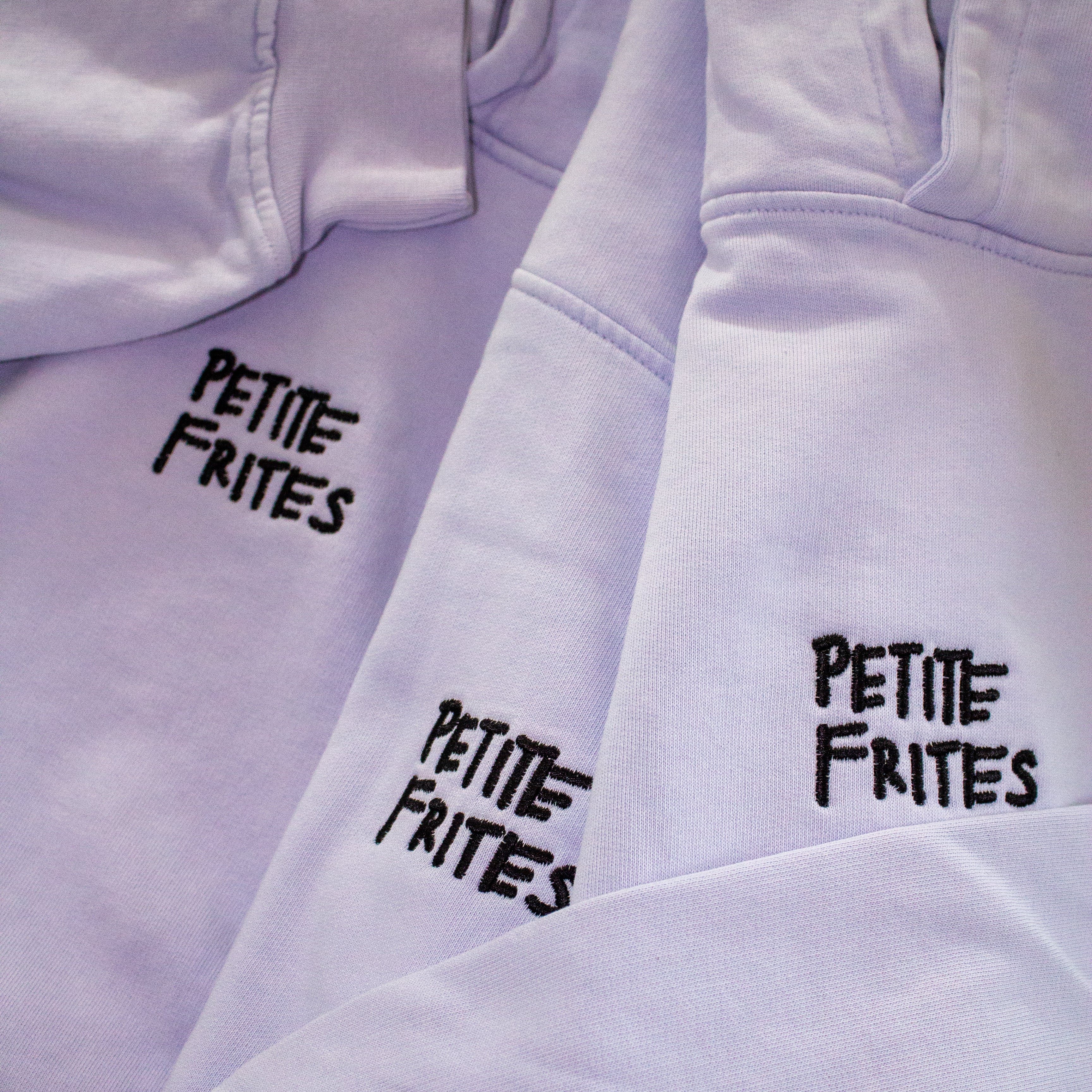 A pile of lavender oversized hoodies with the Petite Frites logo embroidered in black