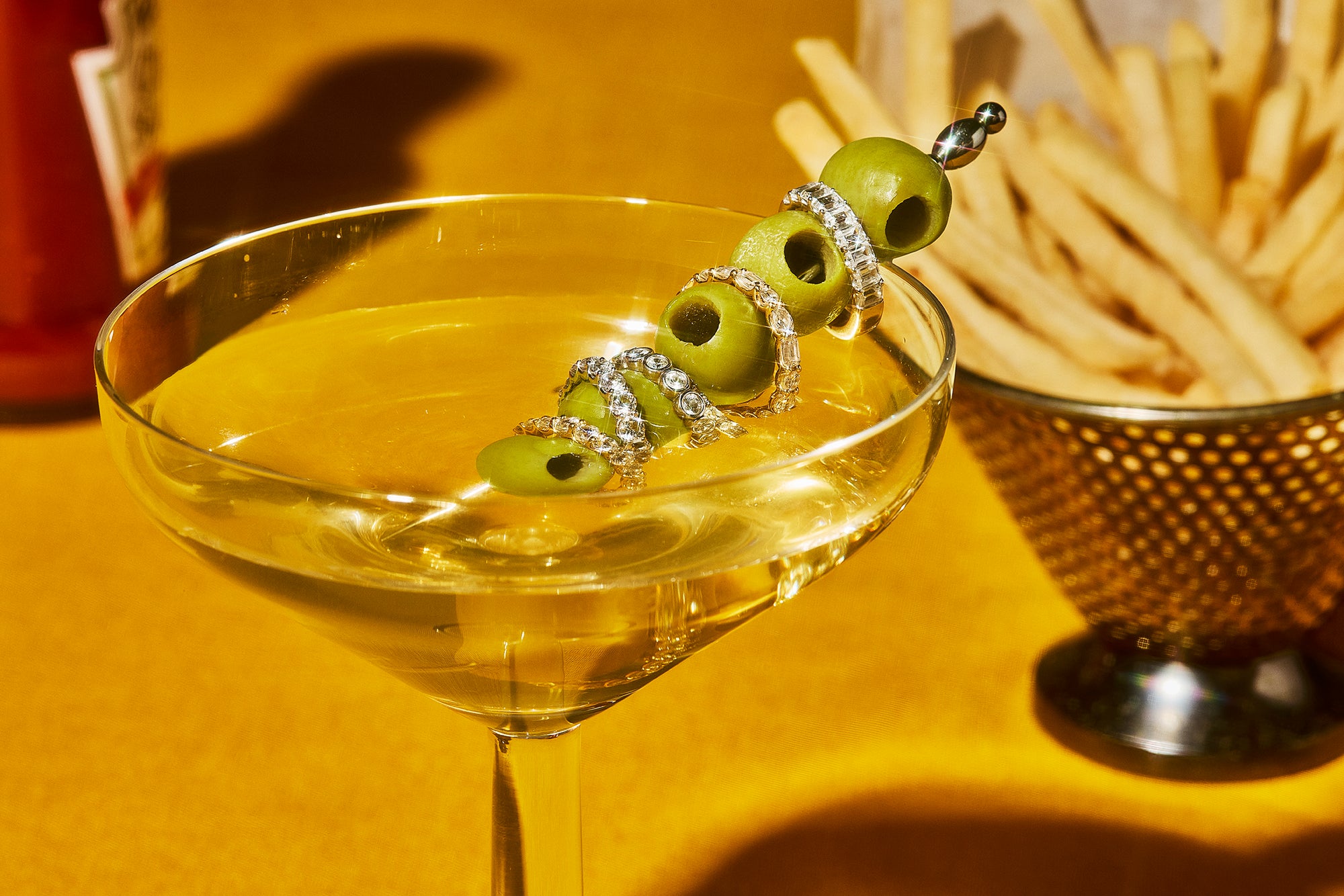 This image shows a martini glass with Petite Frites 18K and lab grown diamond eternity bands stacked between the olives on the martini stick. Behind the glass is a serving of french fries.