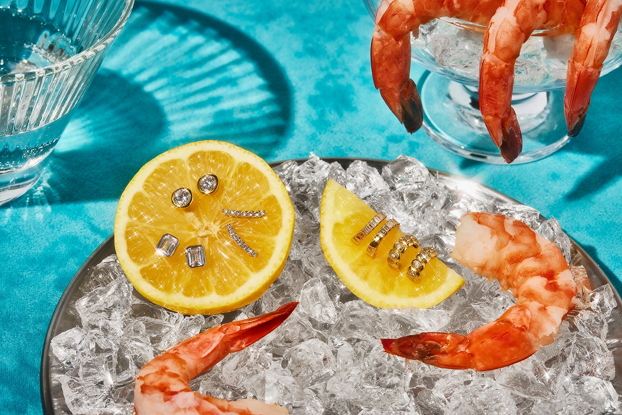 This image shows shrimp cocktail on ice with lemon wedges in a silver platter on a teal tablecloth. On the lemon wedges are Petite Frites 18k gold and carbon neutral lab grown diamond earrings in white and yellow gold. There are studs and hoops. Clicking on this image leads to the earring section.