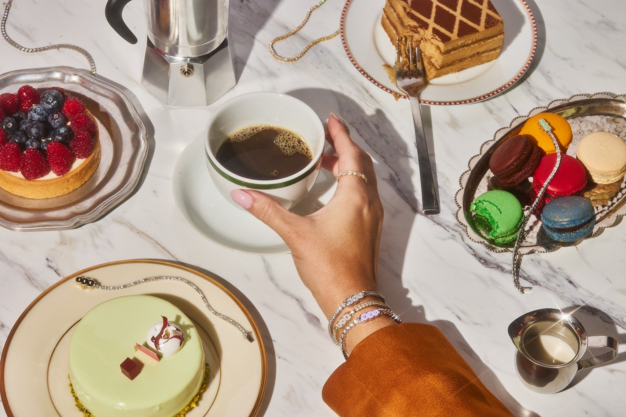 This image shows a female hand wearing 3 Petite Frites 18k gold and carbon neutral lab grown diamond bracelets and a diamond eternity band on the pointer finger. The hand is reaching for a cup of coffee. On the marble table is a dessert buffet of macaroons, a fruit tart, and other colorful cakes, with tennis bracelets placed throughout the display. Clicking on this image leads to the bracelet shop.