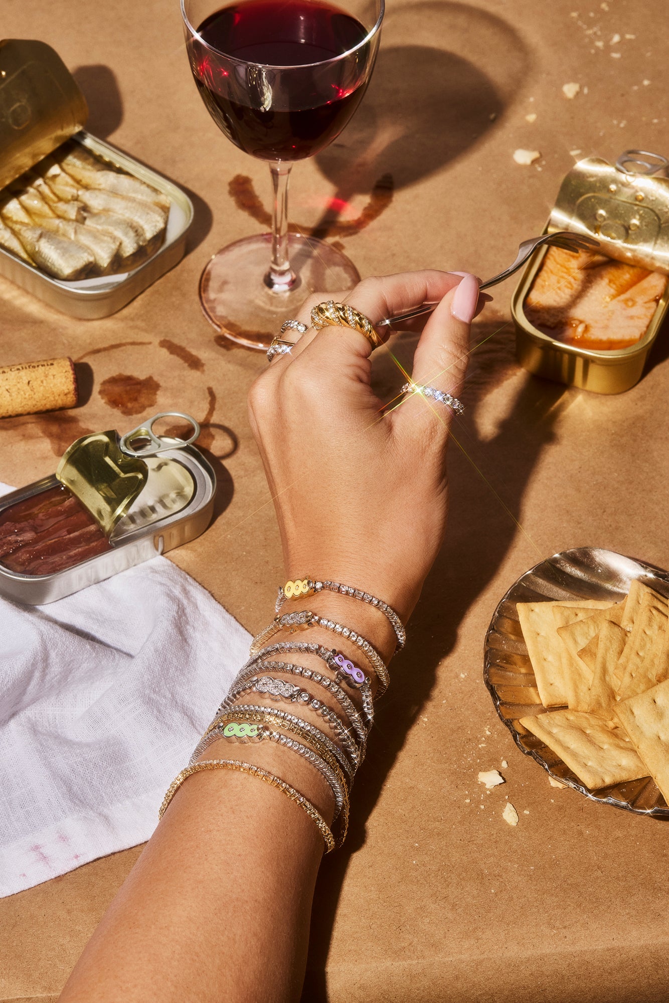This image shows a female hand wearing 8 Petite Frites 18k gold and carbon neutral lab grown diamond tennis bracelets with colorful enamel clasps, including butter yellow, lavender, and lime green. The hand is reaching across a brown table that holds crackers, open sardine cans, and a glass of red wine. On the hand are several Petite Frites rings.