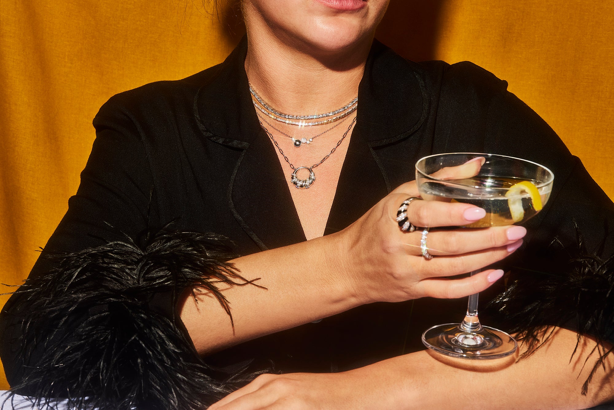 A female in a silky black jacket with black feathers on the sleeves holds a martini glass with her hand that is adorned with Petite Frites 18k gold and carbon neutral lab grown diamond jewelry. She wears several Petite Frites pendants and diamond studded necklaces in white and yellow gold. Clicking on this image leads to the Shop Necklace section.