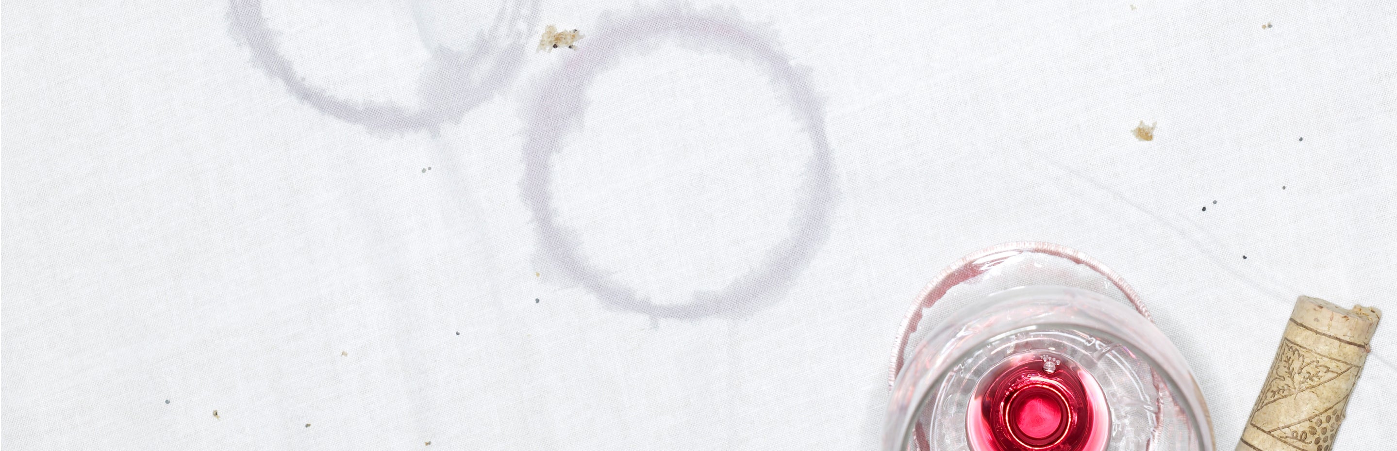 This image shows a white table cloth that has been stained by the bottom of a red wine glass, and a near-empty red wine glass with a cork.