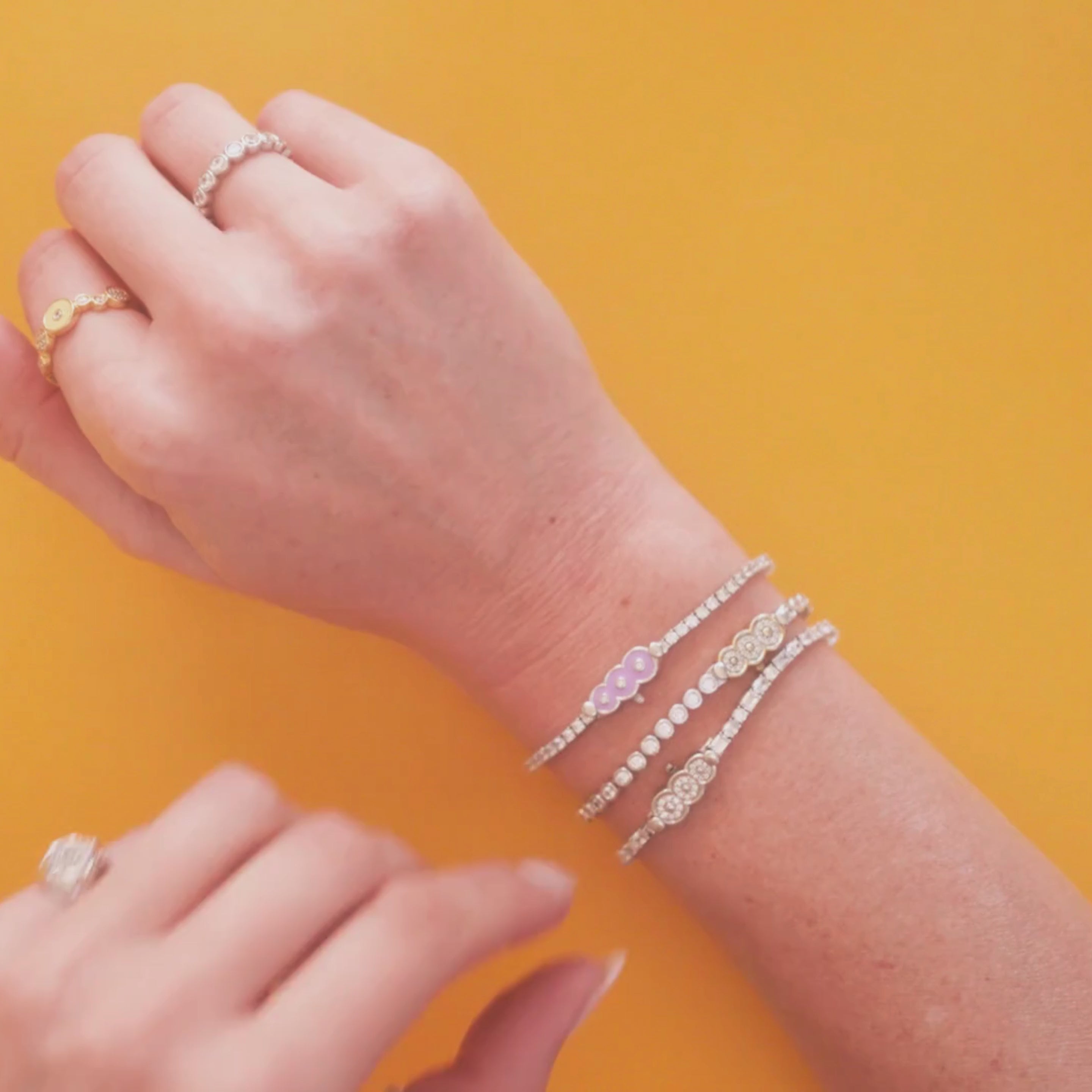 Video of Petite Frites Tennis Bracelets being worn, featuring yellow and white gold diamond clasps, and a lavender enamel clasp on the wrist of a women against a yellow tablecloth.