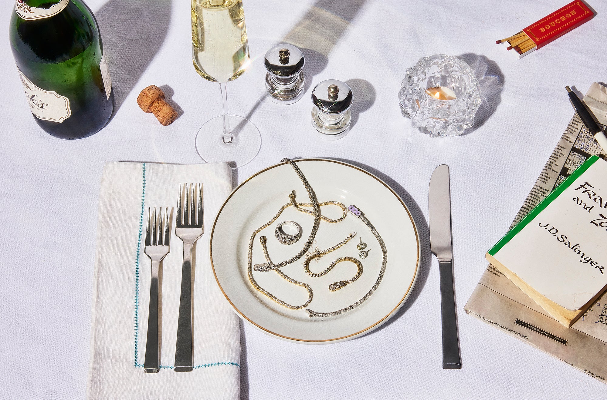 This image shows a dining table with a white linen tablecloth set for 1 with a glass of champagne, candle, crossword puzzle and book. On the plate are Petite Frites 18k gold and carbon neutral lab grown diamond tennis bracelets, earrings, necklaces and rings.