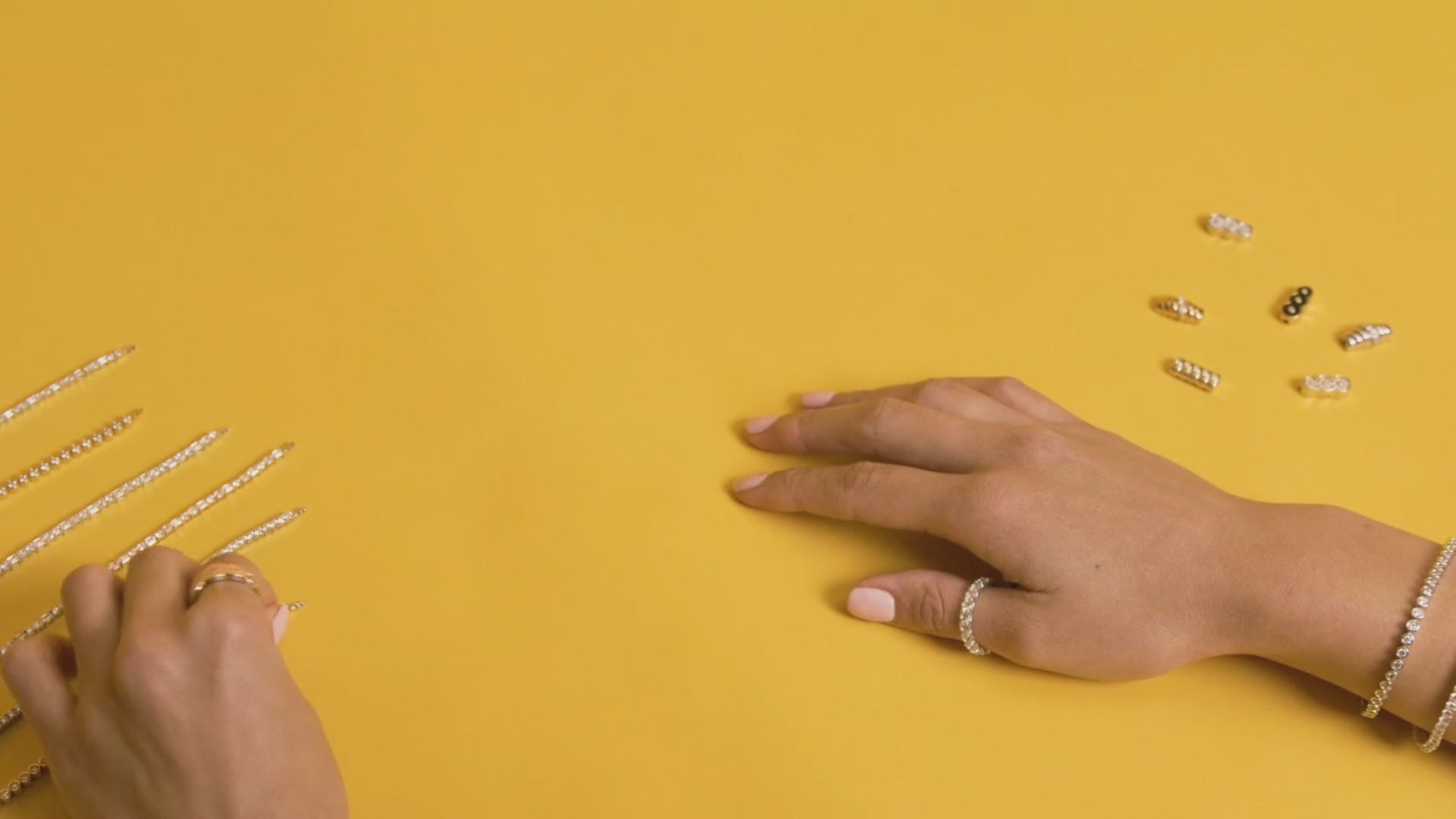Two hands on a yellow table wearing Petite Frites rings and bracelets. The left hand reaches for a bracelet and shows how to put the clasp and bracelet together, and then unlock it.